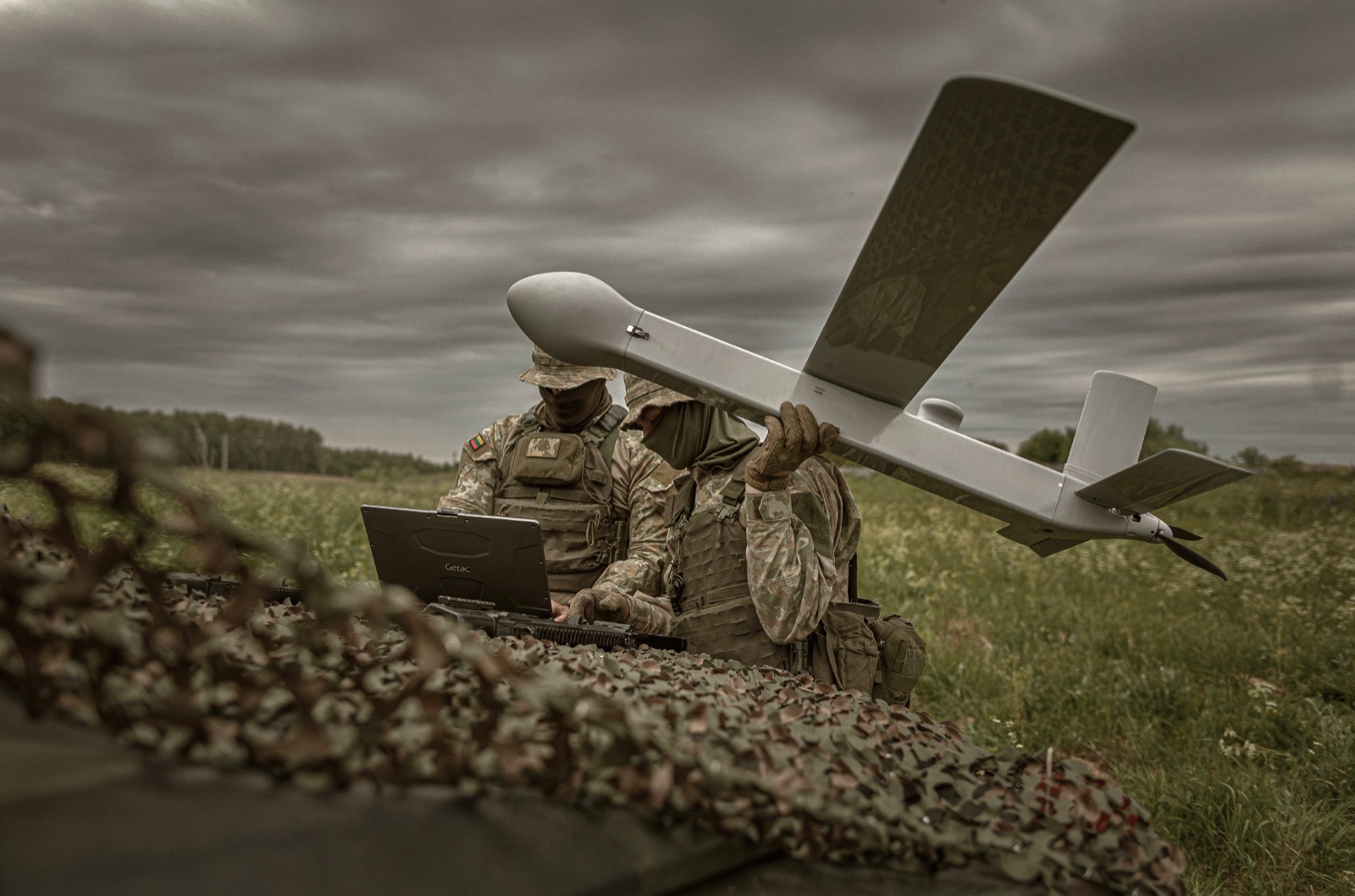 Two operators behind camouflage netting, one at a rugged laptop, the other holding a fixed-wing aircraft ready to launch