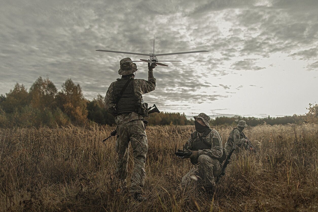 An operator holding a small fixed-wing aircraft overhead before launching it, a second operator kneeling at the controls in long grass